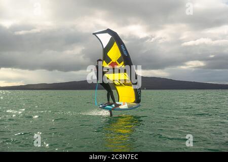 Ein junger Mann wingfoils im Hafen von Auckland, mit einem handaufblasbaren Flügel und Reiten ein Tragflächenboot Surfbrett. Grünes Meer und wolkig blauer Himmel. Stockfoto