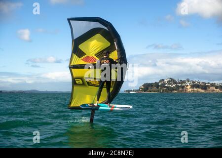 Ein junger Mann wingfoils im Hafen von Auckland, mit einem handaufblasbaren Flügel und Reiten ein Tragflächenboot Surfbrett. Grünes Meer und wolkig blauer Himmel. Stockfoto