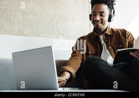 Foto eines lächelnden afroamerikanischen Mannes, der Kopfhörer benutzt, während er mit einem Laptop in einem hellen Raum arbeitet Stockfoto