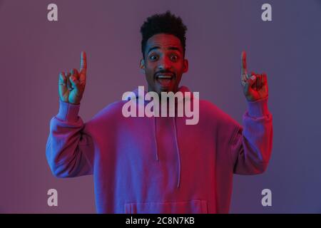 Portrait von spannenden african american man in bunten hoodie auf Kamera und zeigen nach oben isoliert über violette Hintergrund Stockfoto