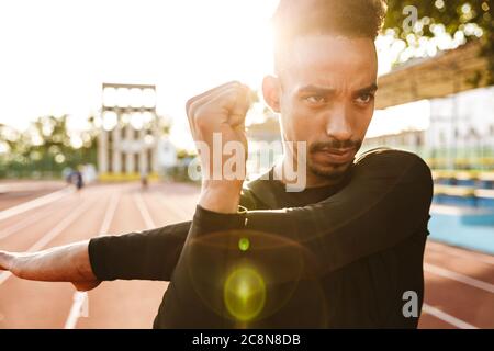 Bild des fokussierten afrikanische amerikanische Mann in Sportswear seine Arme stretching beim Training im Stadion am Morgen im Freien Stockfoto