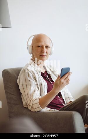 Minimalporträt von kahlköpfigen erwachsenen Frau, die Kamera anschaut, während sie Musik über Smartphone zu Hause hört, Alopezie und Krebsbewusstsein, Kopierraum Stockfoto