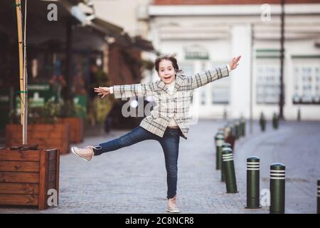 Outdoor-Porträt eines lustigen niedlichen kleinen Mädchen von 8-9 Jahren alt, Wandern und Tanzen auf der Straße, trägt moderne Karos Jacke, Jeans und Turnschuhe Stockfoto