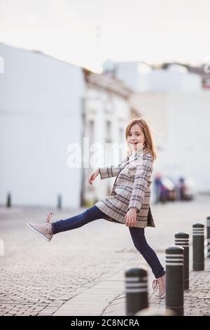 Outdoor-Porträt eines lustigen niedlichen kleinen Mädchen von 8-9 Jahren alt, Wandern und Tanzen auf der Straße, trägt moderne Karos Jacke, Jeans und Turnschuhe Stockfoto