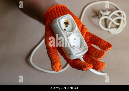 Hand trägt einen orangen Schutzhandschuh hält eine Steckdose nach dem Brand. Weiße Steckdosenleiste nach Kurzschluss. Elektrische Sicherheit. Stockfoto
