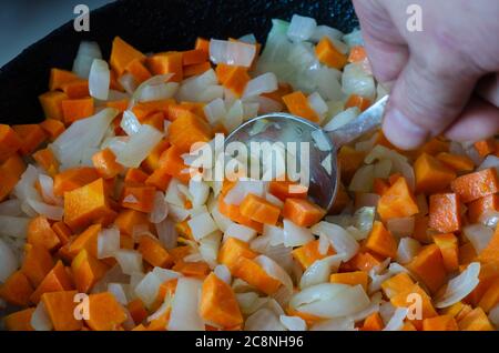 Männliche Hand rührt gewürfelte weiße Zwiebeln und rohe Karotten in der Pfanne. Braten Prozess Scheiben von rohen Zwiebeln und Karotten Nahaufnahme. Draufsicht in einem Winkel. Stockfoto