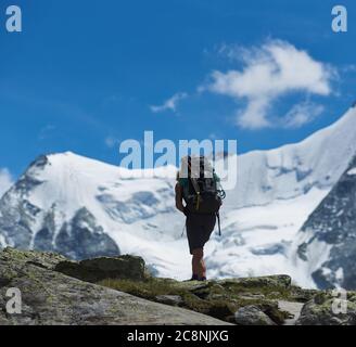 Zinal, Schweiz - 19. Juli 2019: Rückansicht des Reisenden mit Rucksack Wandern allein in der Bergregion. Rucksacktourist, der die Hangstraße mit schneebedeckten Bergen und blauem Himmel im Hintergrund herunterläuft Stockfoto