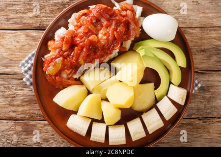 Bermuda-Kabeljau-Frühstück mit Zwiebeltomate-Sauce, gekochten Kartoffeln, Eiern, Banane und Avocado in Nahaufnahme auf einem Teller auf dem Tisch. Horizontale Ansicht von oben her Stockfoto