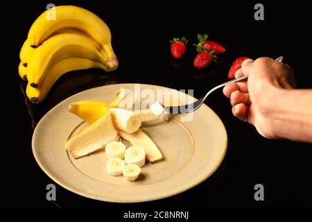 Banane auf einer Keramikplatte Bananenbündel am Boden und eine Hand mit einer Gabel Kommissionierung ein Stück Obst, in schwarzem Hintergrund fotografiert Stockfoto