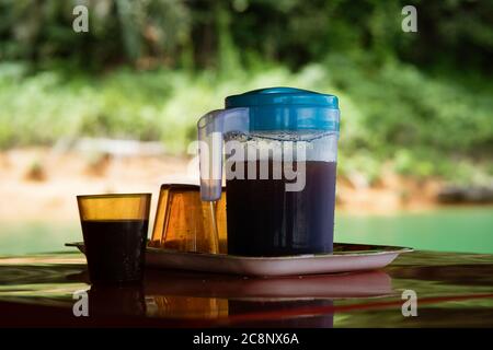 Eiskaffee in einem Plastikbehälter mit leeren Gläsern daneben. Traditionelles kaltes Sommergetränk im Freien in Malaysia. Stockfoto