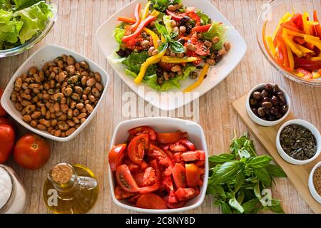 Flach Lay Draufsicht auf eine Verbindung frischen Salatplatte mit pflanzlichen Zutaten rund um ihn auf einem hellen Holztisch und natürliches Sonnenlicht - frische Gesundheit Stockfoto