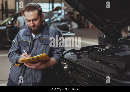 Bärtiger männlicher Mechaniker, der konzentriert auf sein Klemmbrett schrieb und sich auf ein Auto mit offener Kapuze stützte. Erfahrener Autodienstmitarbeiter beim Einfüllen von Papier Stockfoto