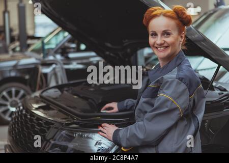 Schöne weibliche Mechanikerin lächelt zur Kamera über ihre Schulter, während sie das kaputte Auto mit einer offenen Kapuze untersucht. Schöne glücklich rothaarige Frau arbeiten Stockfoto