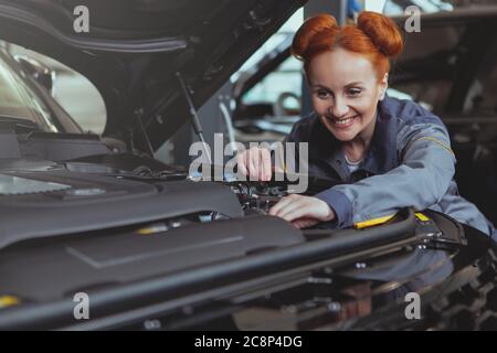 Attraktive fröhliche rothaarige Mechanikerin lächelt freudig und repariert ein Auto in der Garage. Schöne Frau Auto Techniker Reparatur Auto bei Th Stockfoto