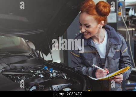 Erfahrene Mechanikerin, die gerne in ihrer Garage arbeitet, Dokumente auf ihrem Klemmbrett ausfüllt und unter die Motorhaube eines Autos schaut. Glückliche schöne Frau Stockfoto
