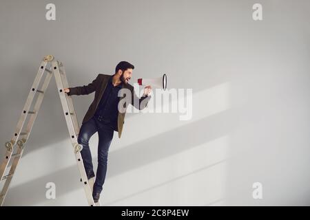 Ein bärtiger Geschäftsmann auf der Treppe mit einem Megafon in der Hand schreit auf grauem Hintergrund. Stockfoto