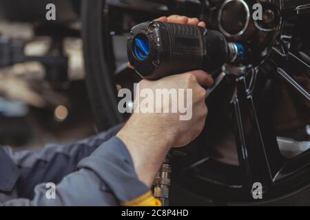 Nahaufnahme eines männlichen Mechanikers in grauer Uniform, der die Anschlagmuttern eines Rades mit einem Elektroschrauber festdreht Stockfoto
