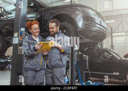 Fröhliche weibliche Mechanikerin im Gespräch mit ihrem männlichen Kollegen, arbeitet an der Autodienststelle, Platz kopieren. Bärtiger Autotechniker, der mit seinem f Stockfoto