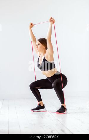 Fit junge Frau tun ein Power-Band hocken Overhead-Widerstand Übung zu stärken und Ton ihre Muskeln in einem High-Key-Fitnessstudio in einem Fitness-Konzept Stockfoto