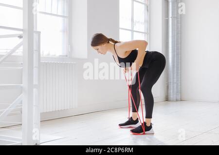 Junge Frau macht Ruderwiderstandübungen mit einem Powerband in einer Seitenansicht in einem High-Key-Fitnessstudio in einem Gesundheits- und Fitnesskonzept Stockfoto