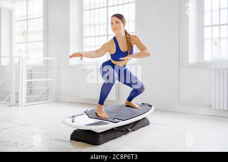 Junge Frau macht ein Surfset regelmäßige Surfer Pose während ihres Fitness-Workout in einem High-Key-Fitnessstudio mit Copyspace in einem gesunden Lifestyle-Konzept Stockfoto