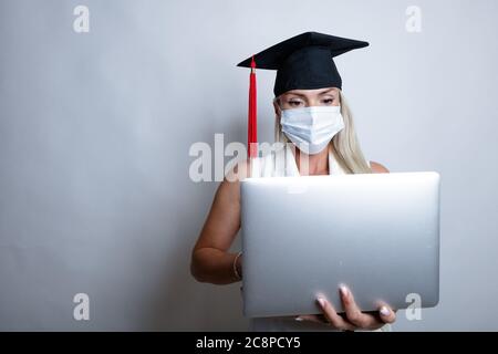 Eine schöne Absolventin in einer Schutzmaske auf ihrem Gesicht und einer Abschlusskappe hält einen Laptop in der Hand. Hochwertige Fotos Stockfoto