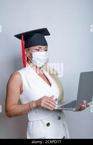 Eine schöne Absolventin in einer Schutzmaske auf ihrem Gesicht und einer Abschlusskappe hält einen Laptop in der Hand. Hochwertige Fotos Stockfoto