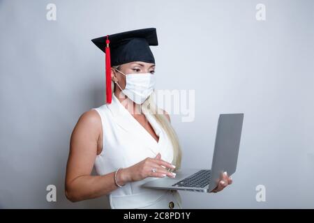 Eine schöne Absolventin in einer Schutzmaske auf ihrem Gesicht und einer Abschlusskappe hält einen Laptop in der Hand. Hochwertige Fotos Stockfoto