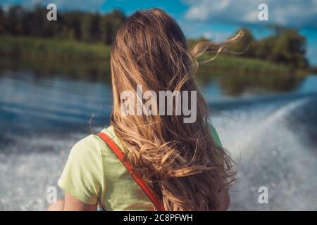 Eine junge Frau mit langen Haaren fährt auf der Wasseroberfläche des Sees mit einem schnellen Boot. Rückansicht. Vor dem Hintergrund einer weißen Schaumstoffspur aus dem Stockfoto