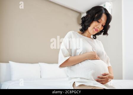 Glücklich schwanger junge asiatische Frau auf dem Bett sitzen und berühren ihren Bauch Stockfoto