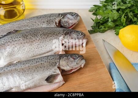 Nahaufnahme von drei rohen Forellenfischen auf einem Holzschneidebrett, das zum Kochen vorbereitet ist. Frische Fischgerichte, gesunde Ernährung und Kochen zu Hause Konzept. Stockfoto