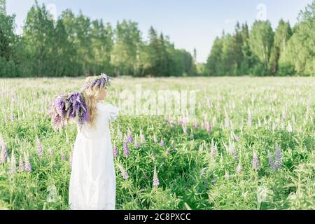 Ein Mädchen mit einem Blumenstrauß über der Schulter in einem weißen Kleid und einem Blumenkranz geht zurück entlang des Blumenfeldes, im Blumenfeld von Stockfoto