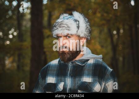 Bärtiger Mann im Winterfell Mütze mit Ohrenklappen Augenschieflage bei der Kamera. Stilvolle männliche tragen kariertes Hemd und Ohrenklappen Pelz Kappe im Herbst fallen Bäume Stockfoto