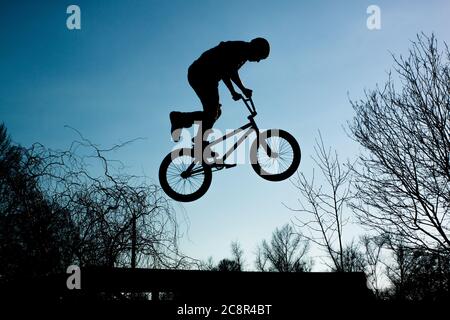 Silhouette eines Mannes, der auf einem Fahrrad gegen einen blauen Himmel mit weißen Wolken springt. Kerl hält Fahrrad Lenker und tun Trick. Stockfoto