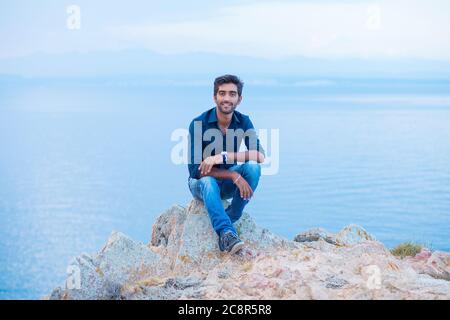 Lächelnder Mann, der die Kamera anschaut, und die Freiheit feiert, während er auf einer Felsklippe über dem Meer sitzt. Positive Person, Peace Mind Konzept. Stockfoto
