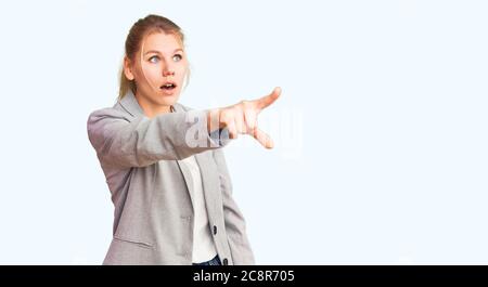 Junge schöne blonde Frau trägt elegante Jacke zeigt mit Finger überrascht voraus, offenen Mund erstaunt Ausdruck, etwas auf der Vorderseite Stockfoto