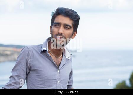 Fröhlicher ethnischer Mann raucht Zigarette, lächelt und denkt, wie Seitenwände draußen vor dem Hintergrund aussehen. Stockfoto