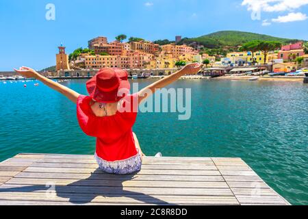 Glückliche Frau, die über dem Steg sitzt und den Hafen von Rio Marina auf der Insel Elba in rotem Anzug betrachtet. Lifestyle Tourist Mädchen auf Urlaubsreise in Italien, Europa Stockfoto