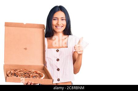 Junge schöne lateinische Mädchen halten Lieferung Pizza Box lächelnd glücklich und positiv, Daumen nach oben tun ausgezeichnet und Genehmigung Zeichen Stockfoto