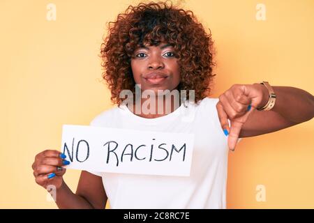 Junge afroamerikanische Frau, die kein Rassismus-Banner mit wütender Gesichtsausdruck hält, negatives Zeichen, das Abneigung mit Daumen nach unten zeigt, Ablehnung Konzept Stockfoto