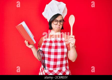 Brunette Frau mit Down-Syndrom tragen professionelle Bäcker Schürze Lesen Kochen Rezept Buch skeptisch und nervös, runzelte wegen des Problems. Stockfoto