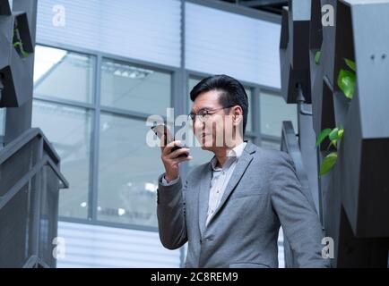 Chinesischer Geschäftsmann trägt eine Jacke, lächelt und benutzt sein Handy, um eine Sprachnachricht zu hinterlassen Stockfoto
