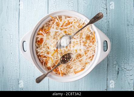 Pfanne voll geriebenem Kohl und Karotte auf blauem Holztisch. Salz und Pfeffer in zwei Metalllöffeln sind auf der Pfanne mit Gemüse. Fermentierter Kohl c Stockfoto