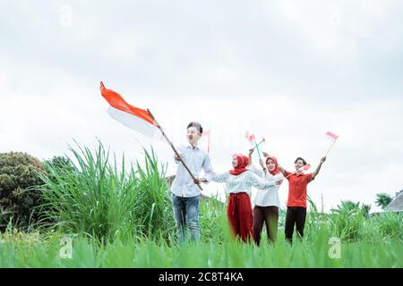 asiatischer junger Mann in Weiß, der eine indonesische Flagge mit Stock trägt und drei seiner Freunde mit einer kleinen Flagge hinter ihm laufen Stockfoto