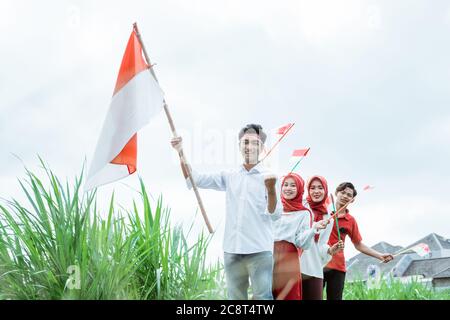 asiatischer junger Mann in weißer Trage hebt eine indonesische Flagge mit Stock und drei seiner Freunde dahinter folgen ihm mit einer kleinen Flagge Stockfoto