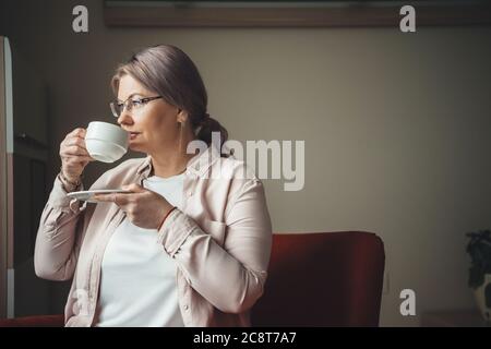 Nachdenkliche kaukasische Frau mit blondem Haar, die einen Kaffee am Fenster trinkt Stockfoto