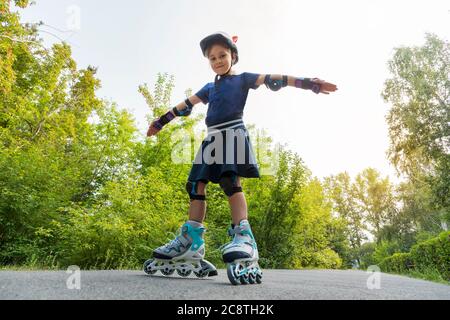 Ein Kind reitet auf Rollschuppen mit ausgestreckten Armen vor dem Hintergrund der grünen Pflanzen im Park. Kleines Mädchen auf Rollschuhe in einem Park Stockfoto
