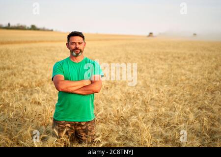 Glücklicher Bauer stolz im Weizenfeld mit gekreuzten Armen auf der Brust. Agronom trägt Corporate Uniform, Blick auf die Kamera auf Ackerland. Reich Stockfoto
