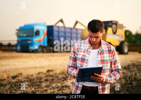 Farmer steuert das Verladen von Weizen vom Erntemaschinen zum Getreidewagen. Fahrer hält Zwischenablage, Notizen, Frachtzählung. Spediteur füllt Sendung aus Stockfoto