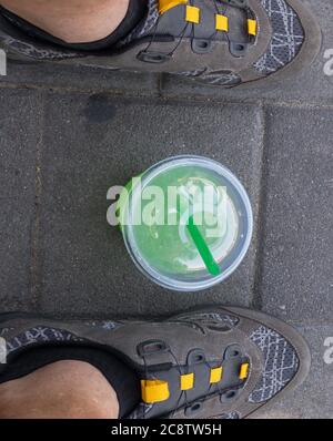 Draufsicht auf Plastikbecher mit grünlicher Flüssigkeit (Mojito) und Cocktail-Rohr innen, stehend auf Bürgersteig zwischen menschlichen Füßen Radschuhe Stockfoto